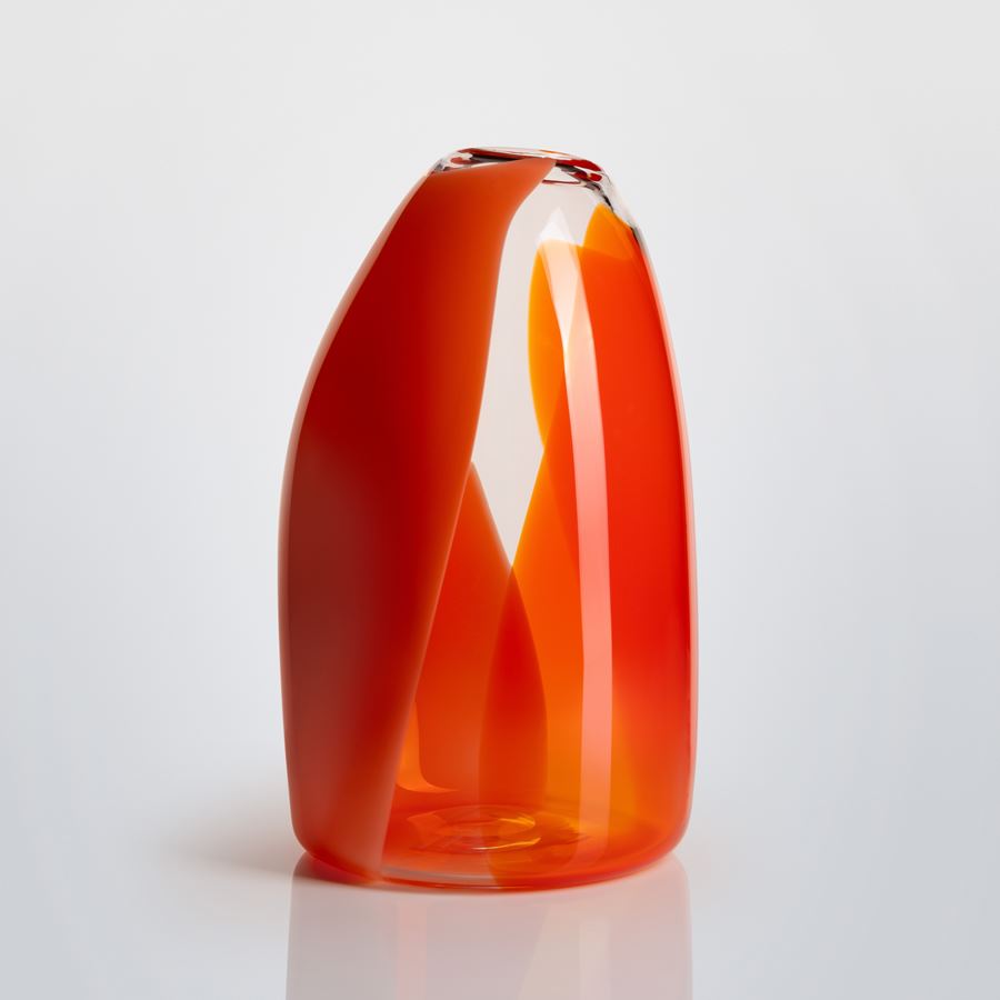 curved narrowing at the top tall transparent glass vase with soft hazy bands of colour in orange red coral and pink curling round the form hand made and blown from glass 
