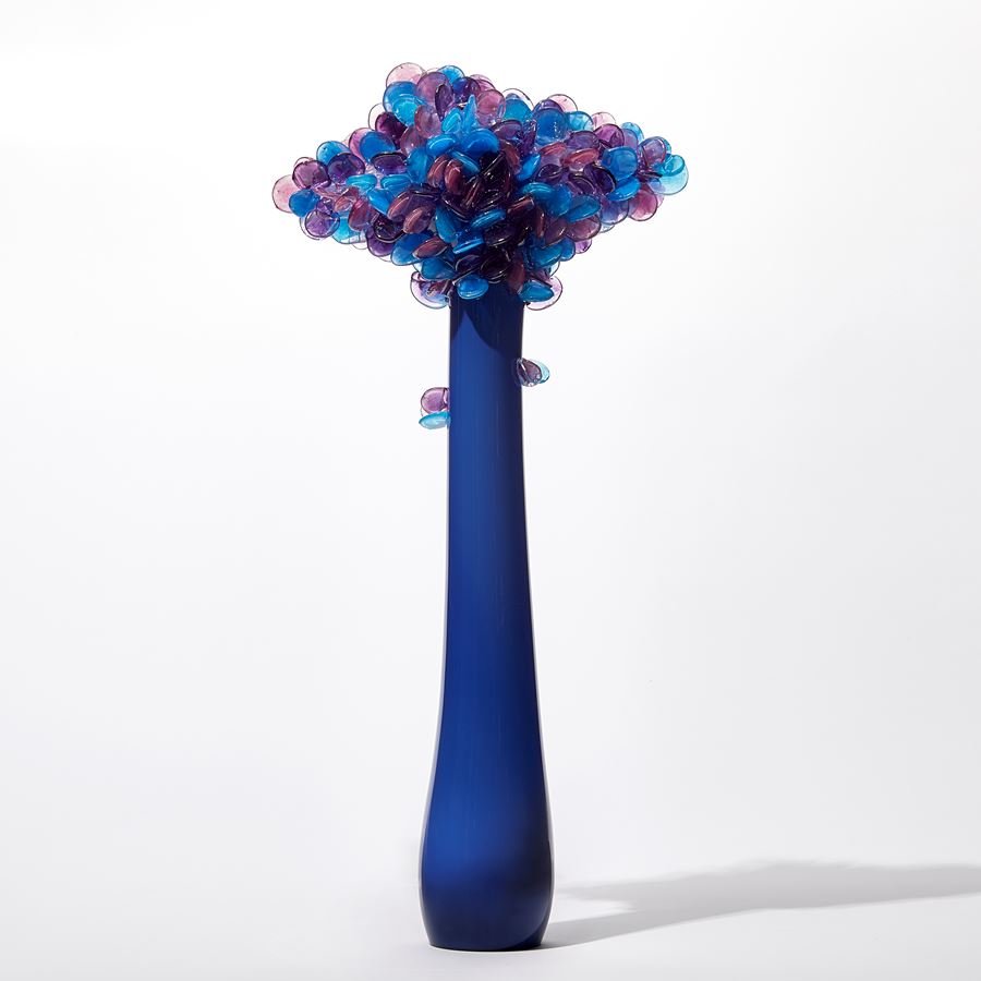 tall tree sculpture with long elegant thin trunk in opaque royal blue with diamond shaped canopy covered in lollipop shaped leaves in blue purple and lilac hand made from glass
