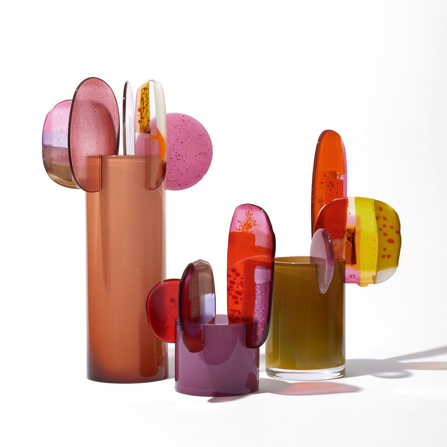 opaque khaki short tubular vase with three rounded finials in differing sizes overlapping and perched on the top rim in pink brown red gold yellow and clear hand made from blown and fused glass