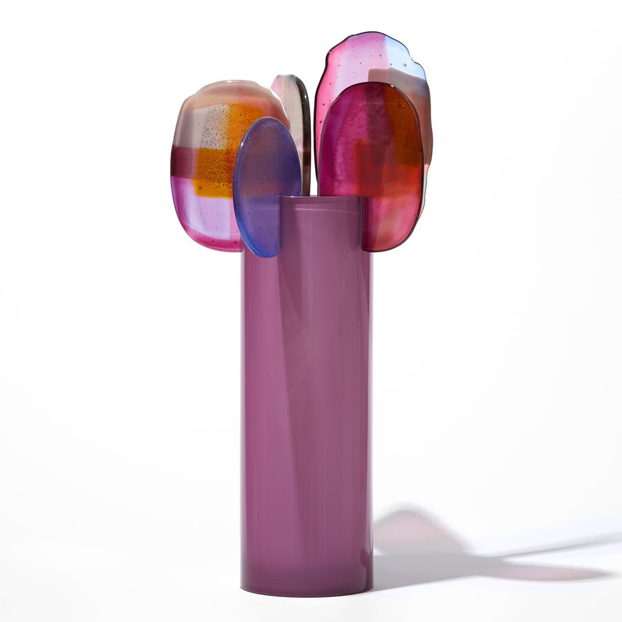 opaque raspberry pink cylinder with five rounded finals with abstract patterns in pink blue purple and gold perched on the top edge hand made from blown and fused glass