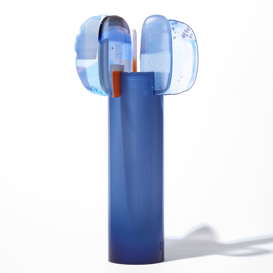 cornflower blue cylinder with five rounded abstract petal forms perched on top with patches of colour in red blue lilac and yellow hand made from blown and fused glass
