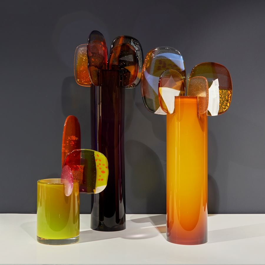 tall opaque shiny rich brown coloured cylinder with 5 rounded finials perched on the top rim in a dazzling array of colours pink yellow red orange and grey with abstract dots and patterns hand made from glass