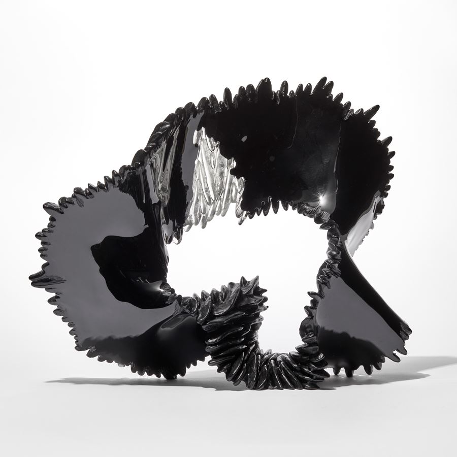 black ridged and textured standing glass sculpture with the appearance of a loop unravelling ribbon with one small clear upper wedged section