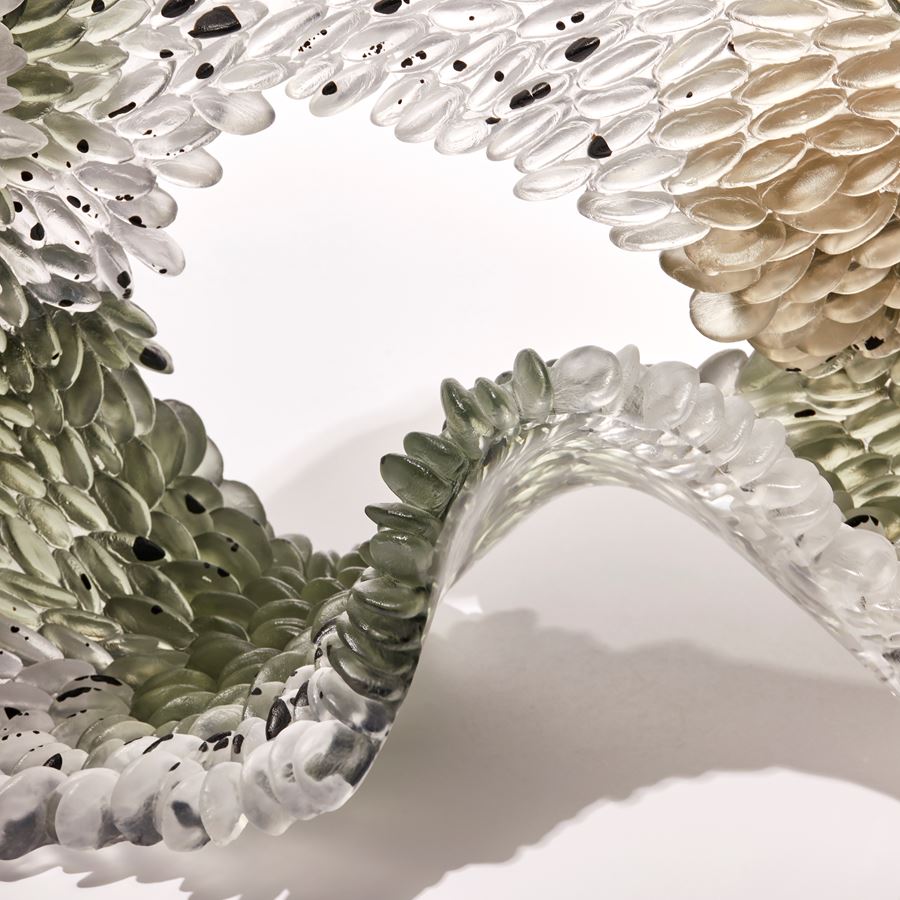 clear textured organically looped standing glass sculpture with one side scaled the other smooth with areas in clear soft moss green and bronze with speckles and dots of opaque black