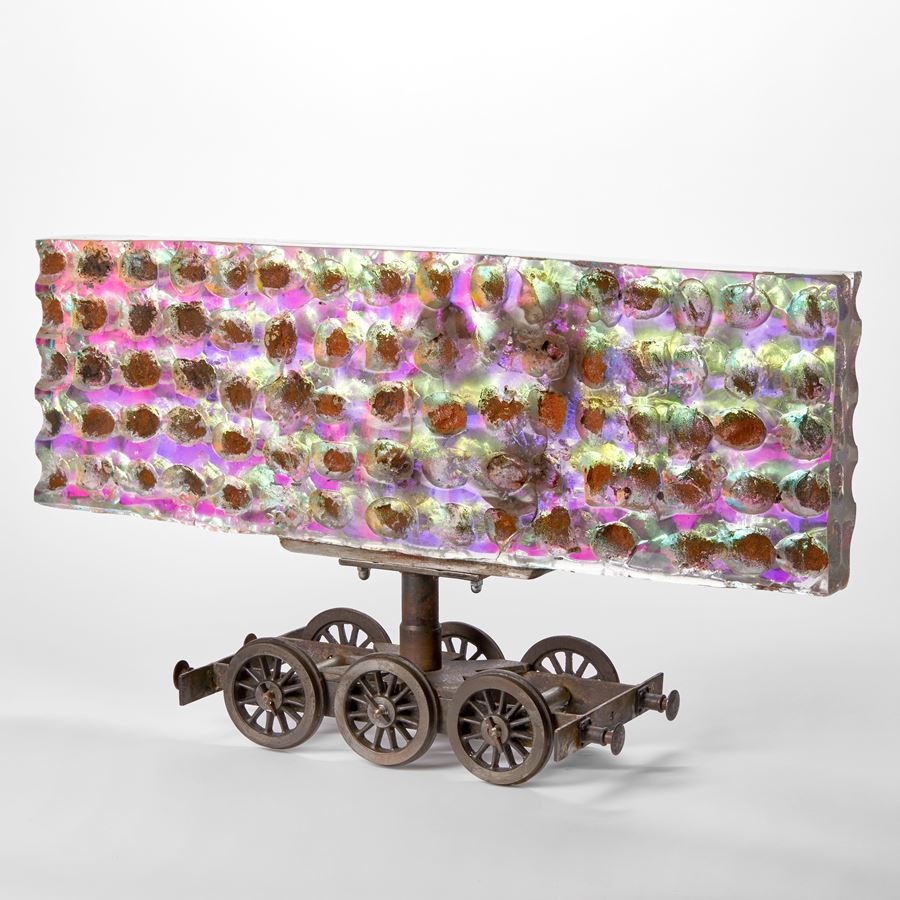 steel six wheel train chassis with a large rectangular bill board shaped sheet of thick cast glass above with sandwiched filter in the centre creating a shimmering and emitting a spectrum of colours in blue greens lilacs and purples with a rough dimpled surface with rusted wells