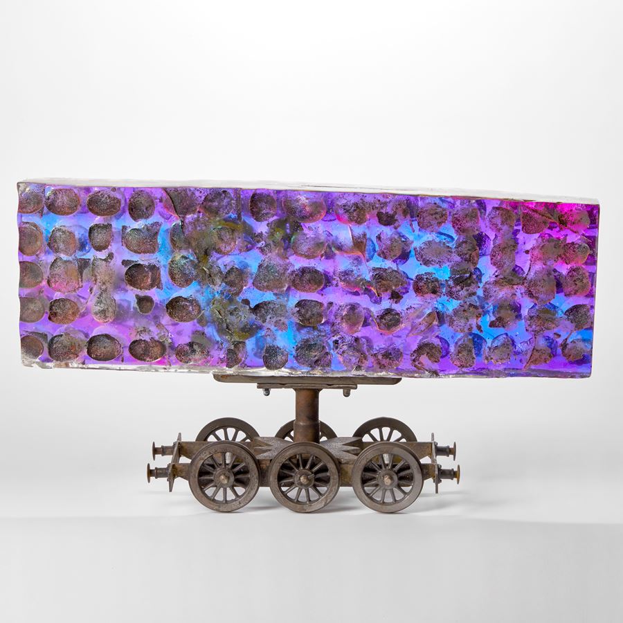 steel six wheel train chassis with a large rectangular bill board shaped sheet of thick cast glass above with sandwiched filter in the centre creating a shimmering and emitting a spectrum of colours in blue greens lilacs and purples with a rough dimpled surface with rusted wells
