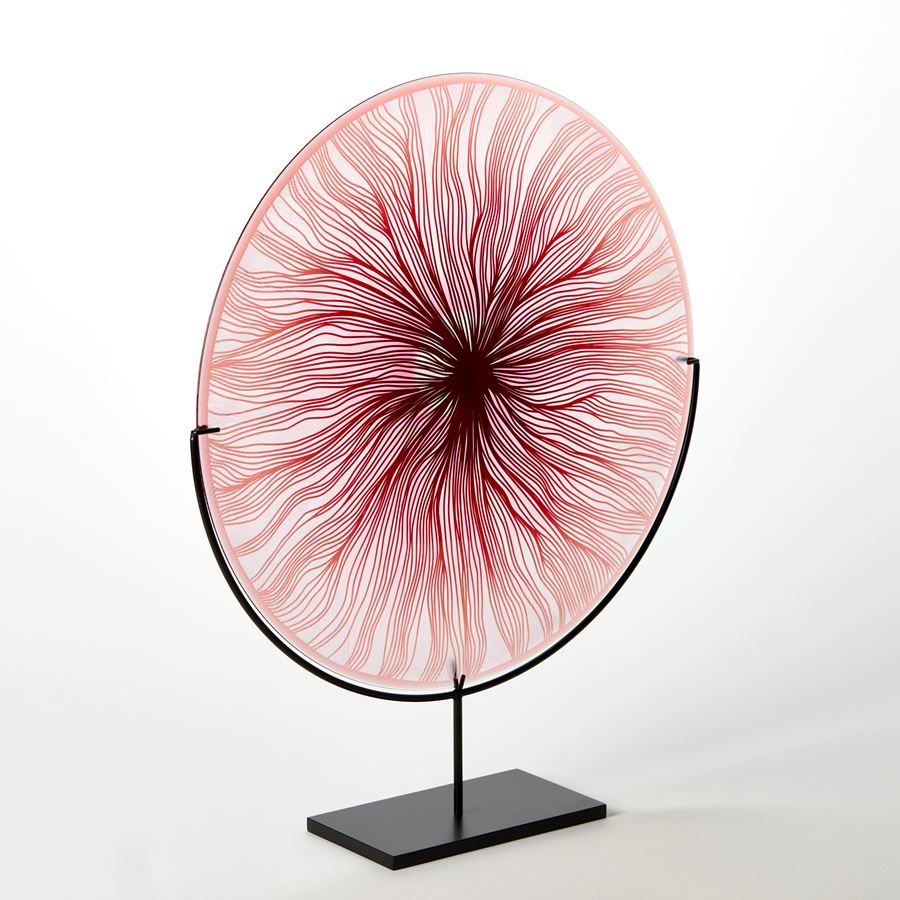 round glass rondel in red pink and clear with cut wavy lines emanating from the centre hand made from glass and presented on a matt black steel stand