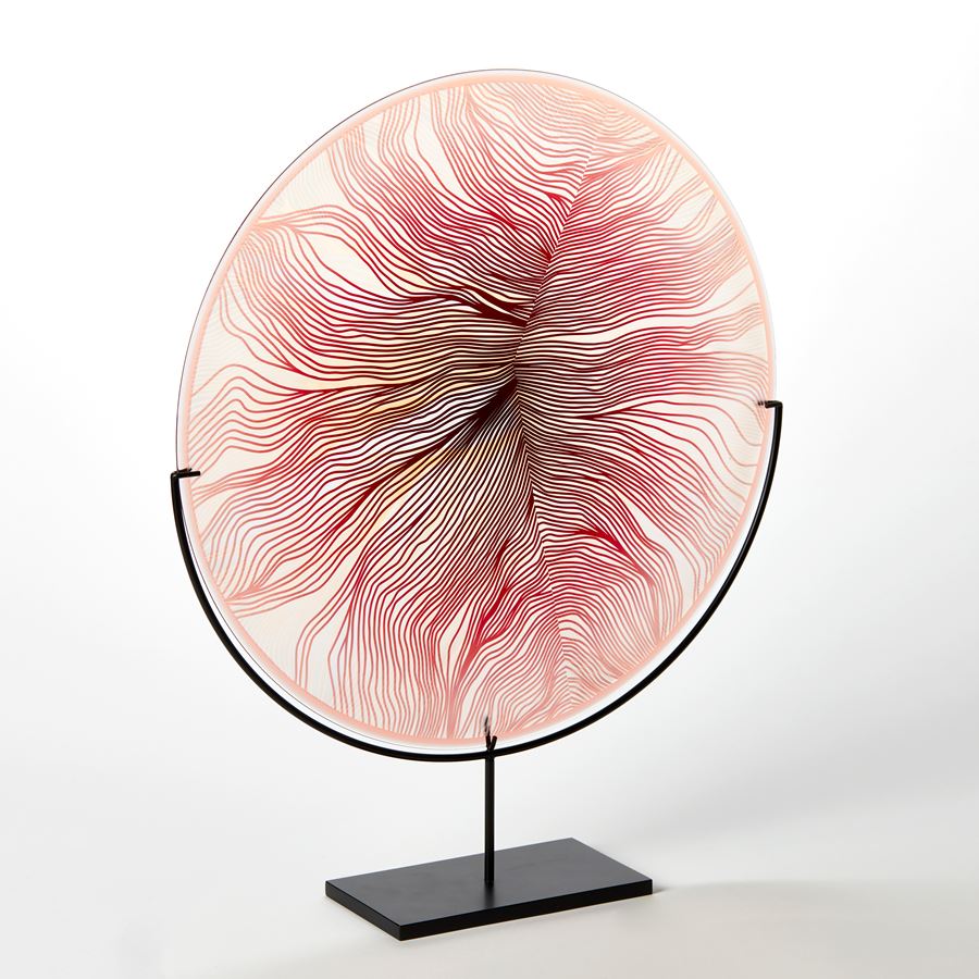 round glass rondel in red pink and transparent glass with surface cut pattern with the appearance of part of a feather hand made from glass on a matt black steel stand