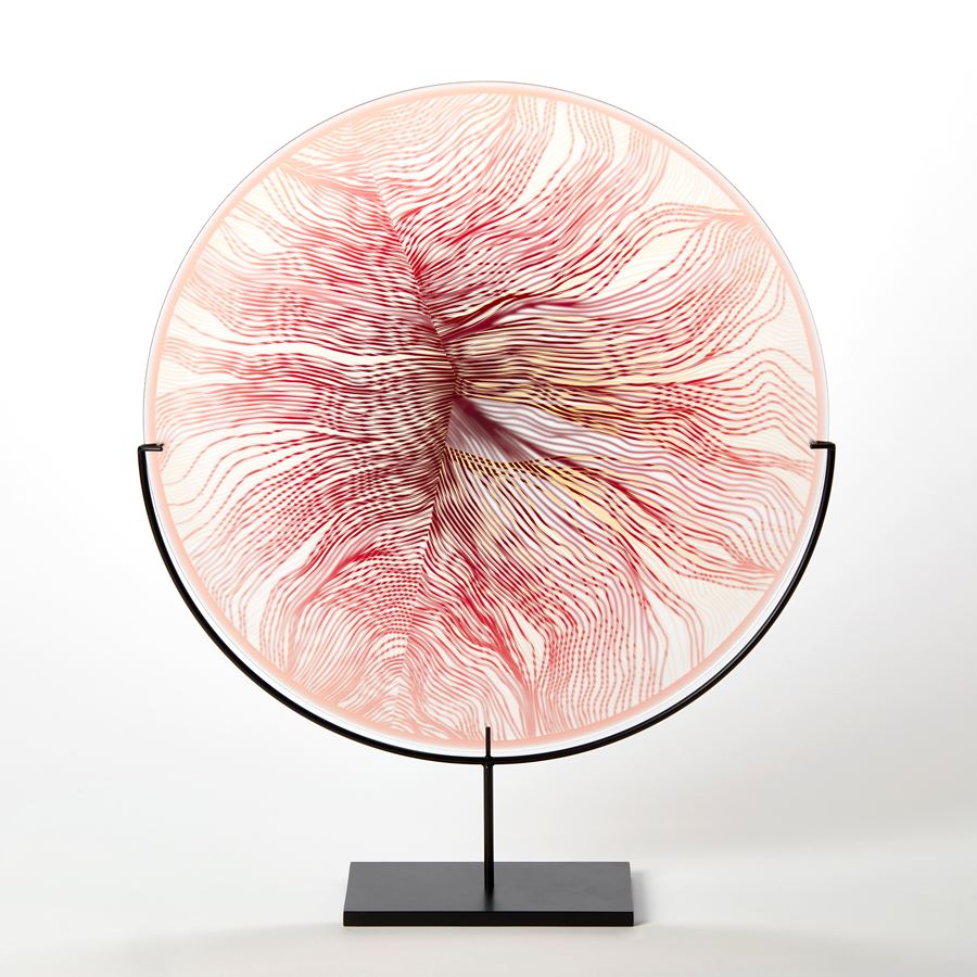 round glass rondel in red pink and transparent glass with surface cut pattern with the appearance of part of a feather hand made from glass on a matt black steel stand