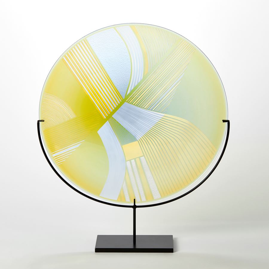 round glass rondel in yellow lime and dove grey blue with abstract landscape patterns cut on the surface handmade from glass and held aloft on a black matt stand