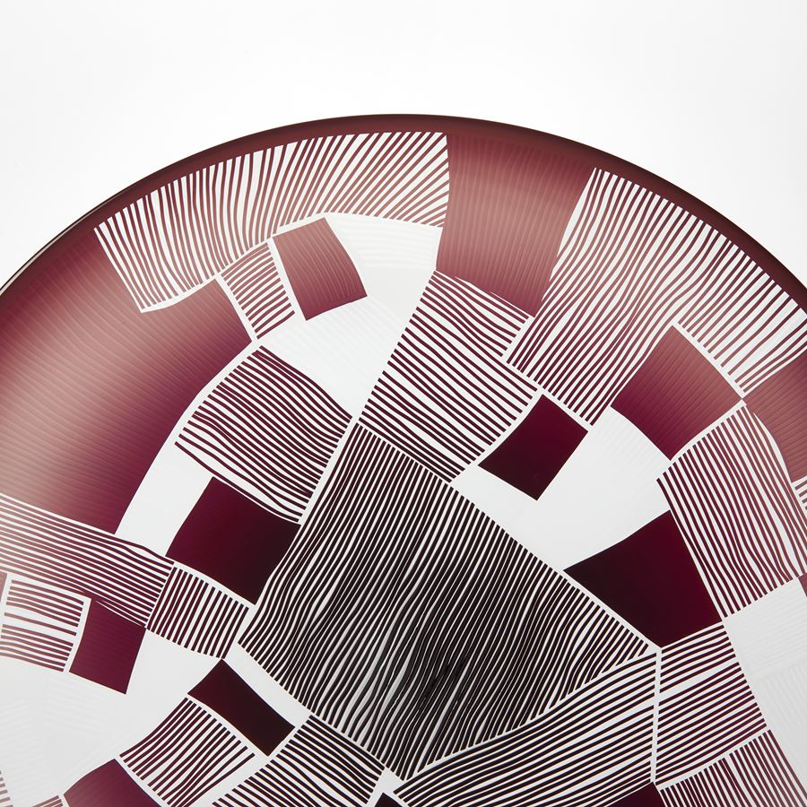 clear and oxblood red round glass plate with patchwork landscape abstract cutting on the surface presented on a matt black steel stand