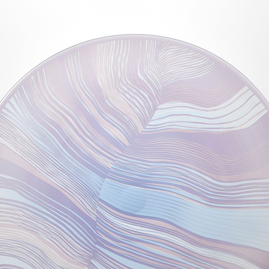 round translucent rondel in soft lilac and aqua with abstract cut lines on the surface similar to a birds feather handblown from glass presented on a matt black stand