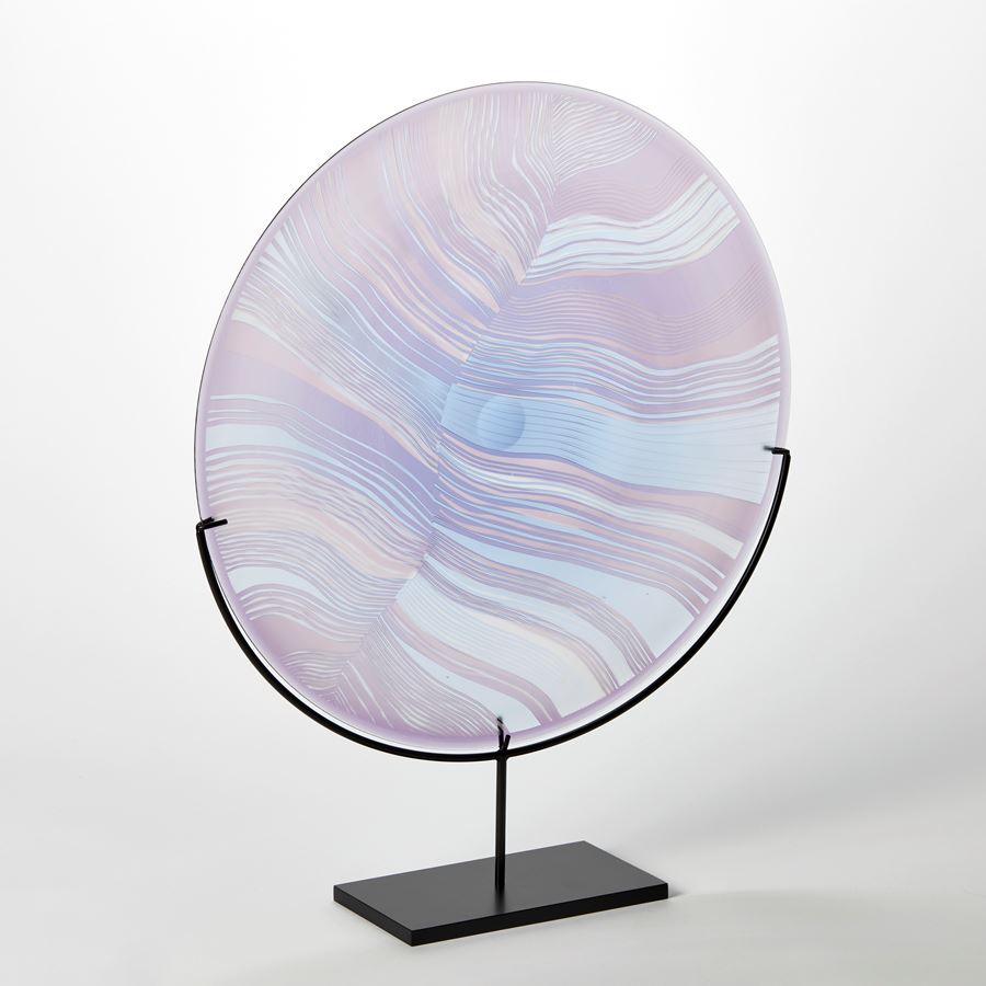 round translucent rondel in soft lilac and aqua with abstract cut lines on the surface similar to a birds feather handblown from glass presented on a matt black stand
