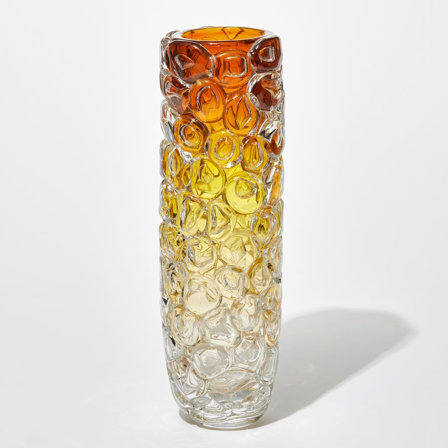 tall round column shaped transparent vase with the outer surface covered in oversized squashed bubbles with clear base fading to soft gold then rich gold and dark amber at the top hand made from glass