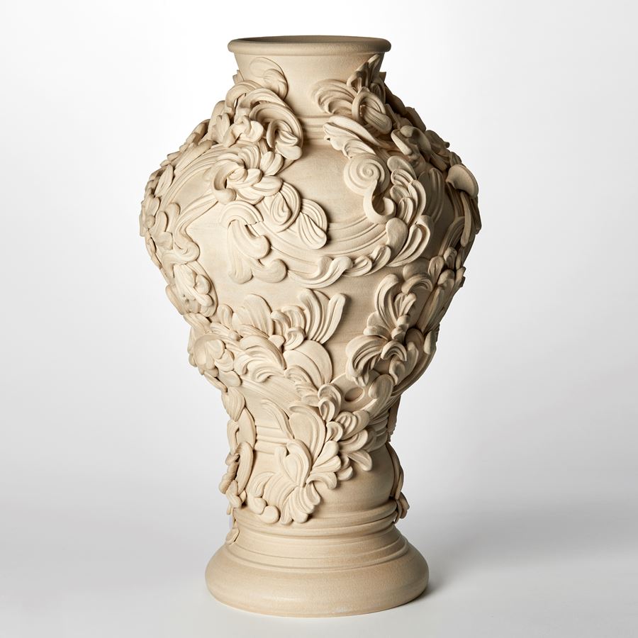 tall classical sandstone coloured vase with narrowing base bulging upper middle and top narrower round opening covered with architectural rococo swirls and flourishes hand made from white st thomas clay  