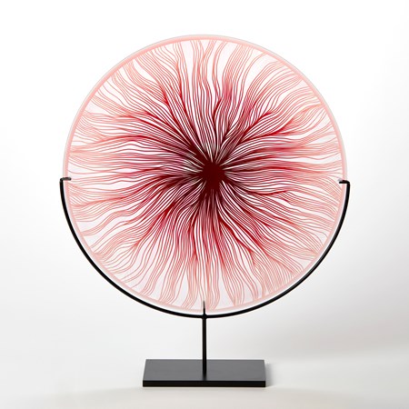round glass rondel in red pink and clear with cut wavy lines emanating from the centre hand made from glass and presented on a matt black steel stand