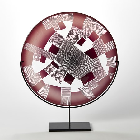 clear and oxblood red round glass plate with patchwork landscape abstract cutting on the surface presented on a matt black steel stand