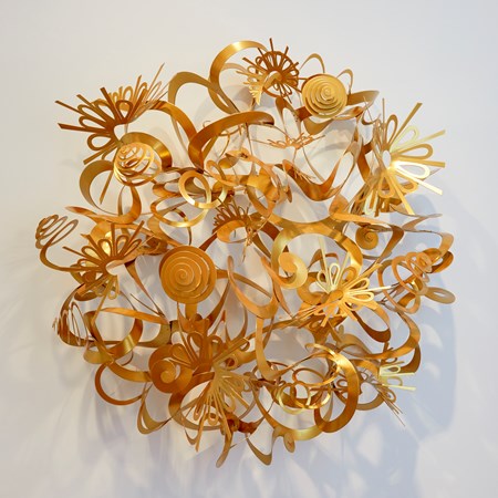round domed wall mounted sculpture with a matt golden finish representing the sun consisting of ribbons swirls curls and shapes filling the negative space