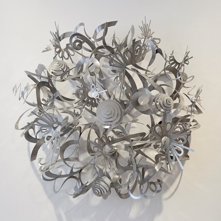 wall mounted round artwork consisting of a mass of swirls curls springs ribbons and shapes creating a domed mass all from thin sheet metal and finished in a matt grey silver colour