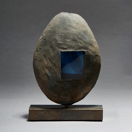 grey and brown hand made glass and metal ovoid shaped sculpture with square glass blue window and square geometric aged metal base
