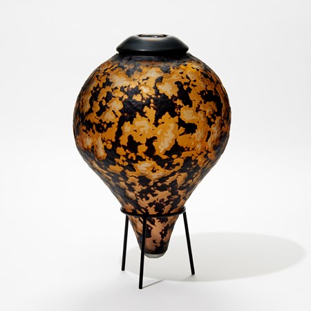 rounded bottle with pointed bottom in black and amber with cut organic textured surface sat on a tripod black steel stand