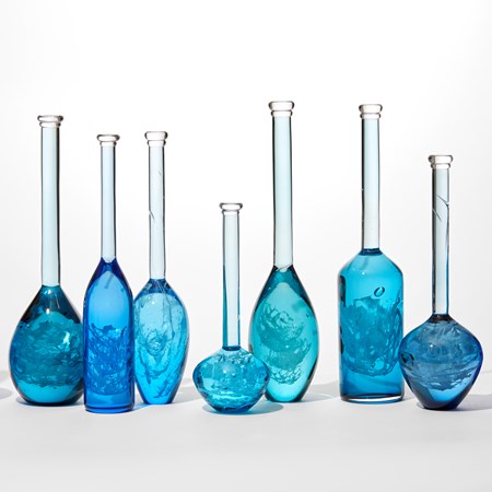 installation of 10 solid bottles in varying hues of jade royal blue aquamarine and turquoise with the suggestion of swirling mysterious contents held within each one all having long thin necks and caps on the top hand blown from glass