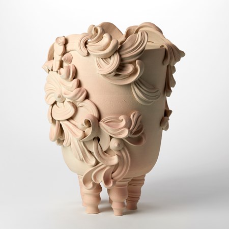bucket shaped biscuit coloured vessel perched on four twisting narrow short legs covered in rounded discs and architectural flourishes with a soft peach blush highlighting some areas hand made from white st thomas clay