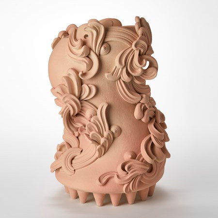 bloated s shaped sculptural vessel in dusty peach pink and beige held aloft on a multitude of small cone shaped feet on the base covered in architectural swirls and curls hand made from white st thomas clay