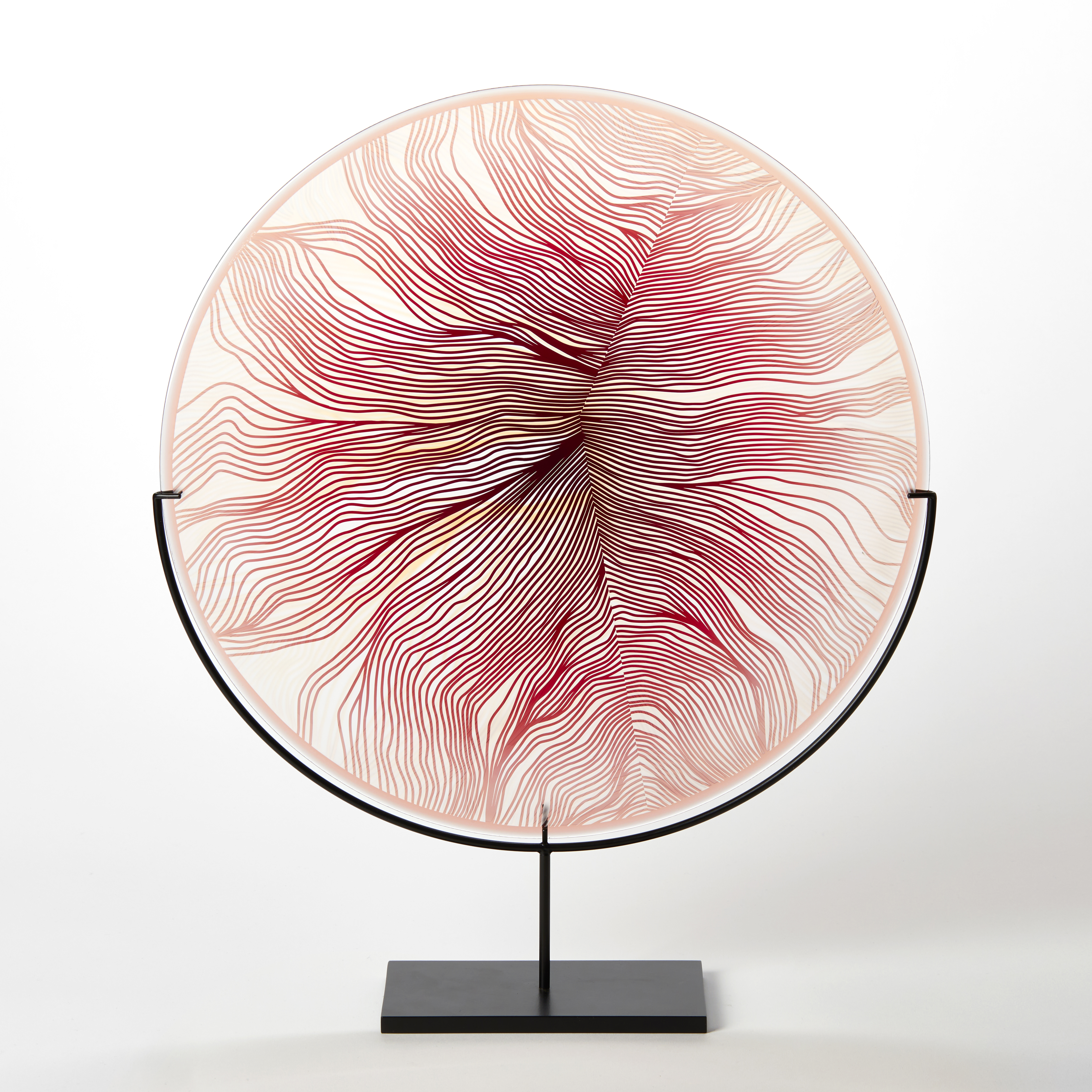 round glass rondel in red pink and transparent glass with surface cut pattern with the appearance of part of a feather hand made from glass on a matt black steel stand