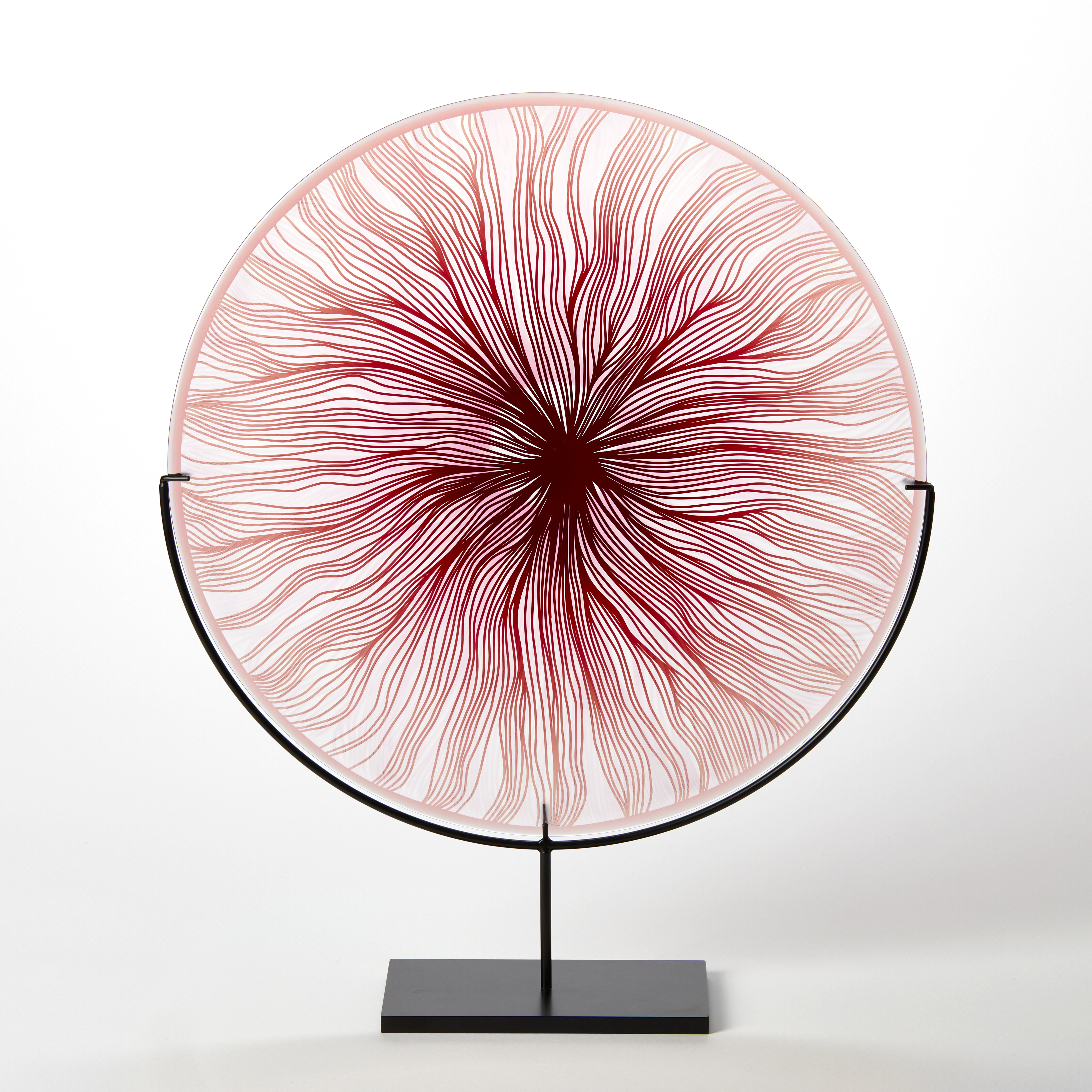 round glass rondel in red pink and clear with cut wavy lines emanating from the centre hand made from glass and presented on a matt black steel stand
