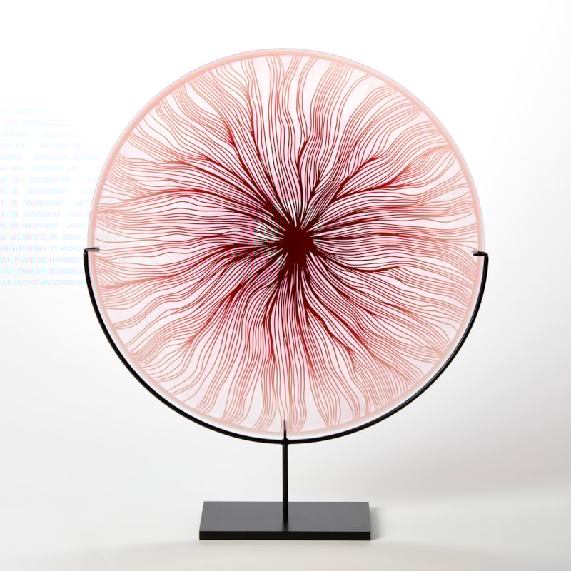 round glass rondel in red pink and clear with cut wavy lines emanating from the centre hand made from glass and presented on a matt black steel stand