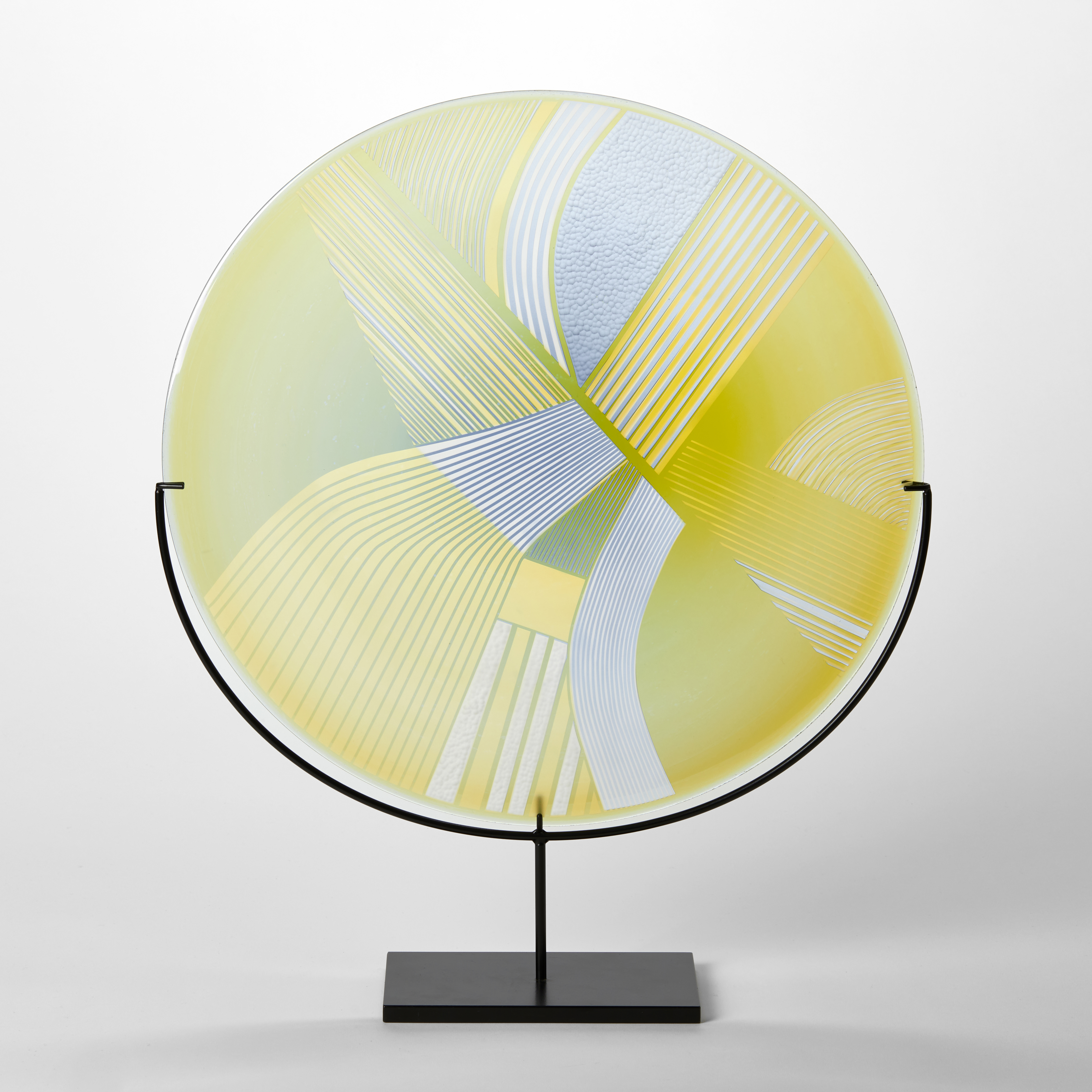 round glass rondel in yellow lime and dove grey blue with abstract landscape patterns cut on the surface handmade from glass and held aloft on a black matt stand