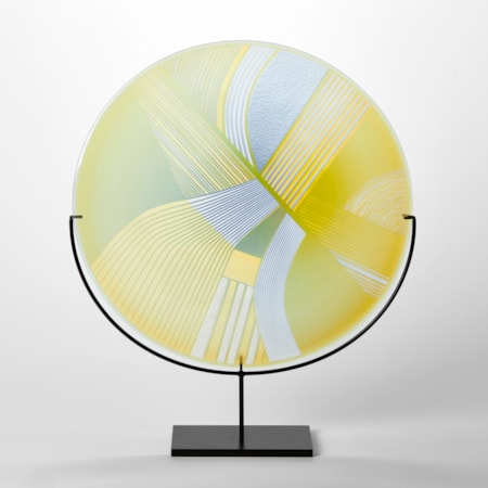 round glass rondel in yellow lime and dove grey blue with abstract landscape patterns cut on the surface handmade from glass and held aloft on a black matt stand