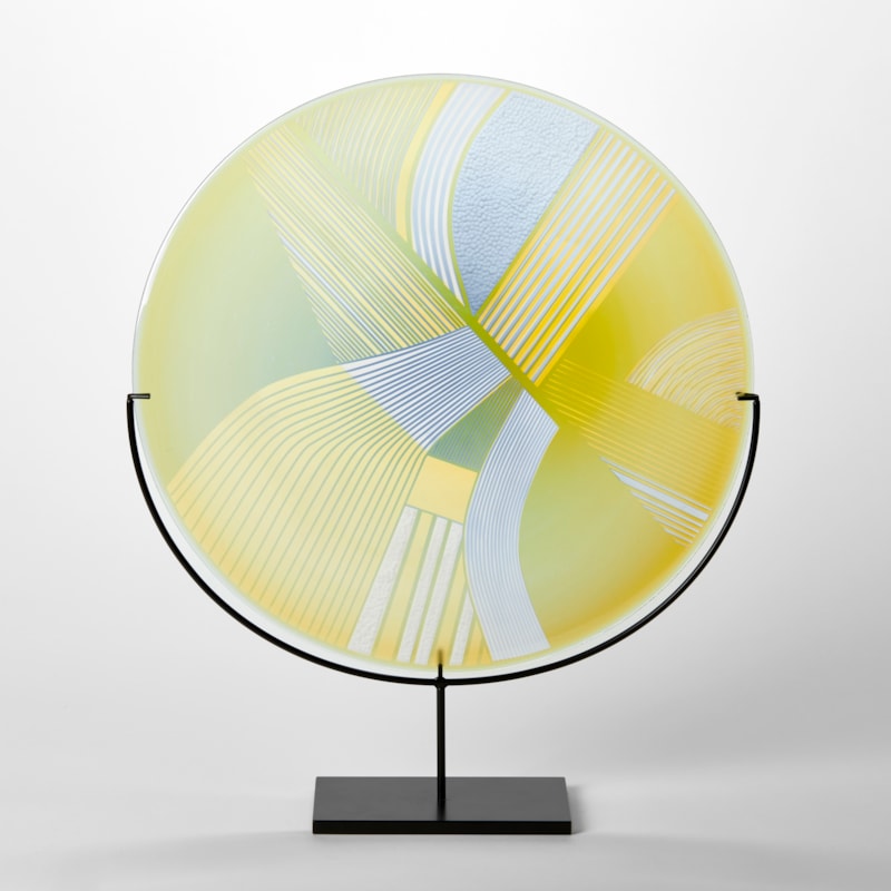 round glass rondel in yellow lime and dove grey blue with abstract landscape patterns cut on the surface handmade from glass and held aloft on a black matt stand