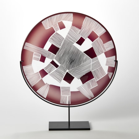 clear and oxblood red round glass plate with patchwork landscape abstract cutting on the surface presented on a matt black steel stand