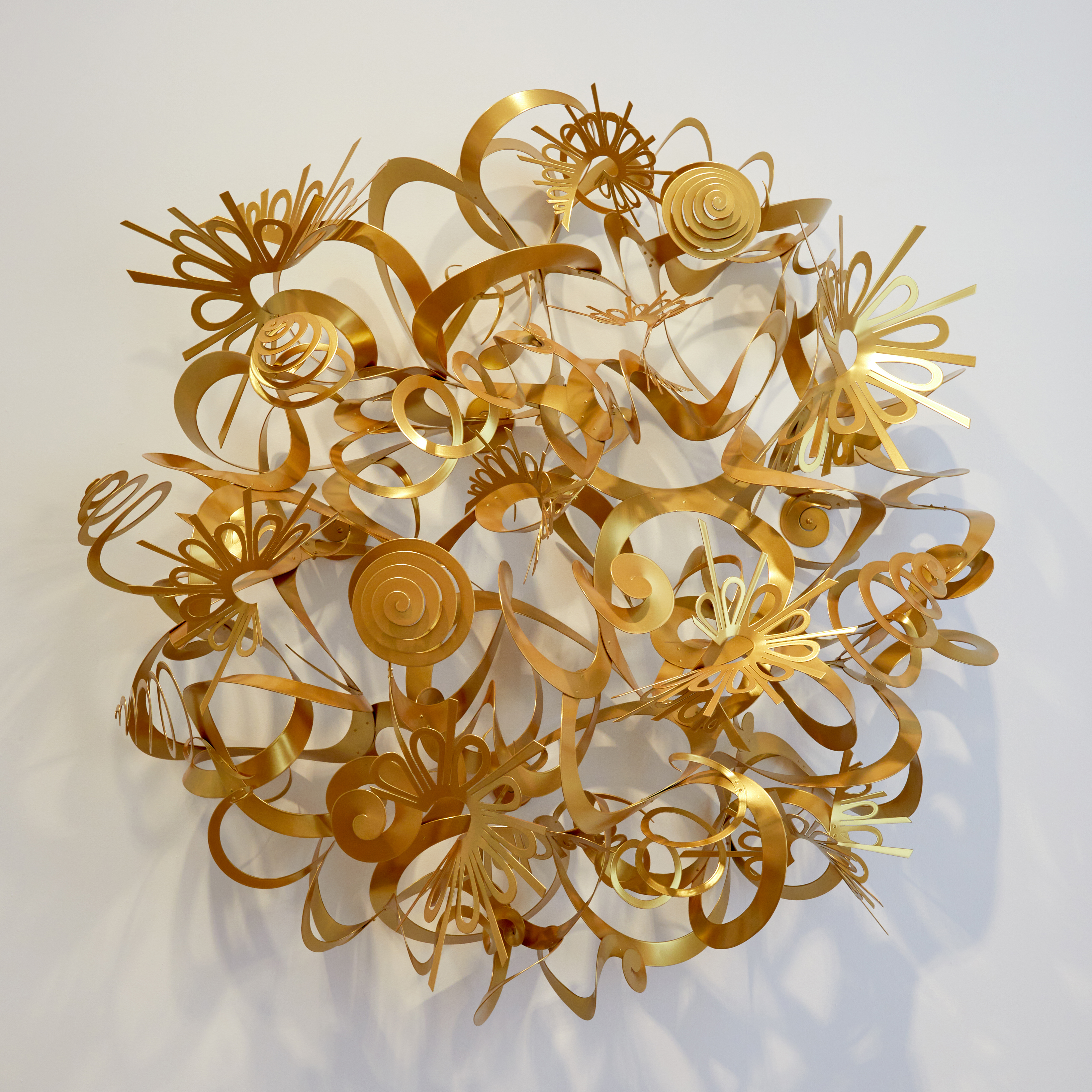 round domed wall mounted sculpture with a matt golden finish representing the sun consisting of ribbons swirls curls and shapes filling the negative space
