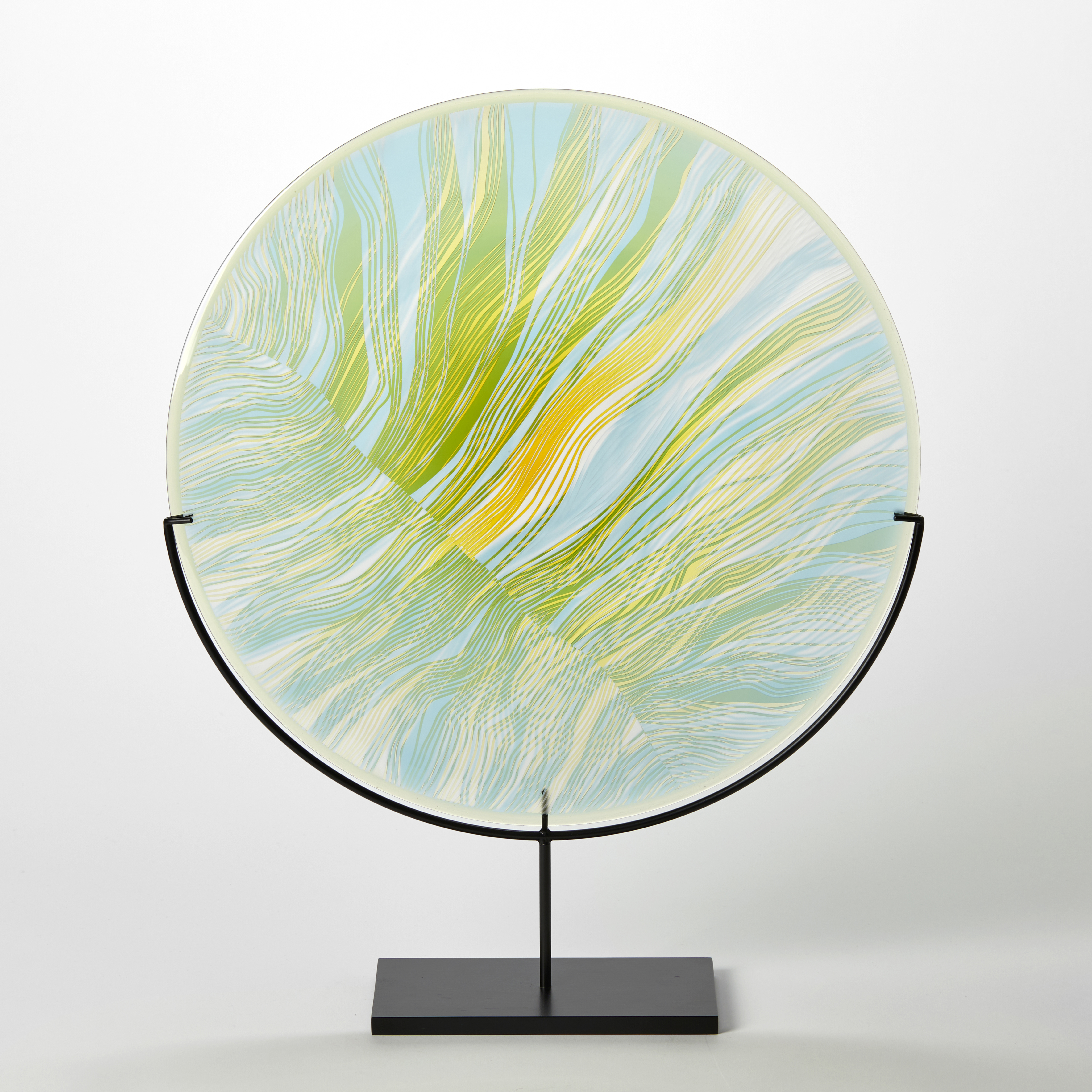 translucent green yellow and aqua round disc on a matt black stand with abstract cut lines on the surface hand made from glass