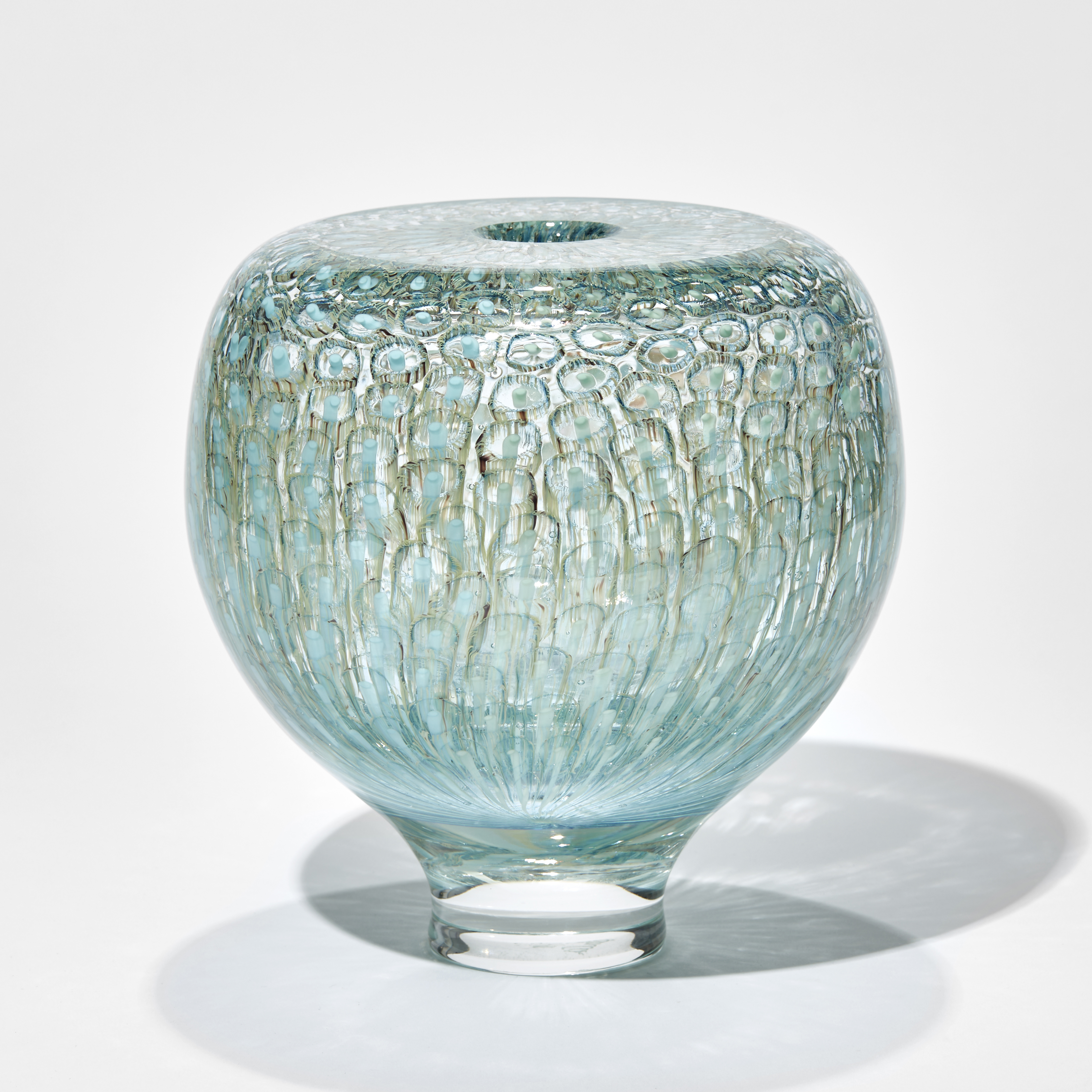 rounded transparent vessel with tapered foot and a flat top with small opening hand made from thick handblown glass with a repeat pattern like tiny abstract jelly fish trapped under the surface in celadon olive teal and turquoise