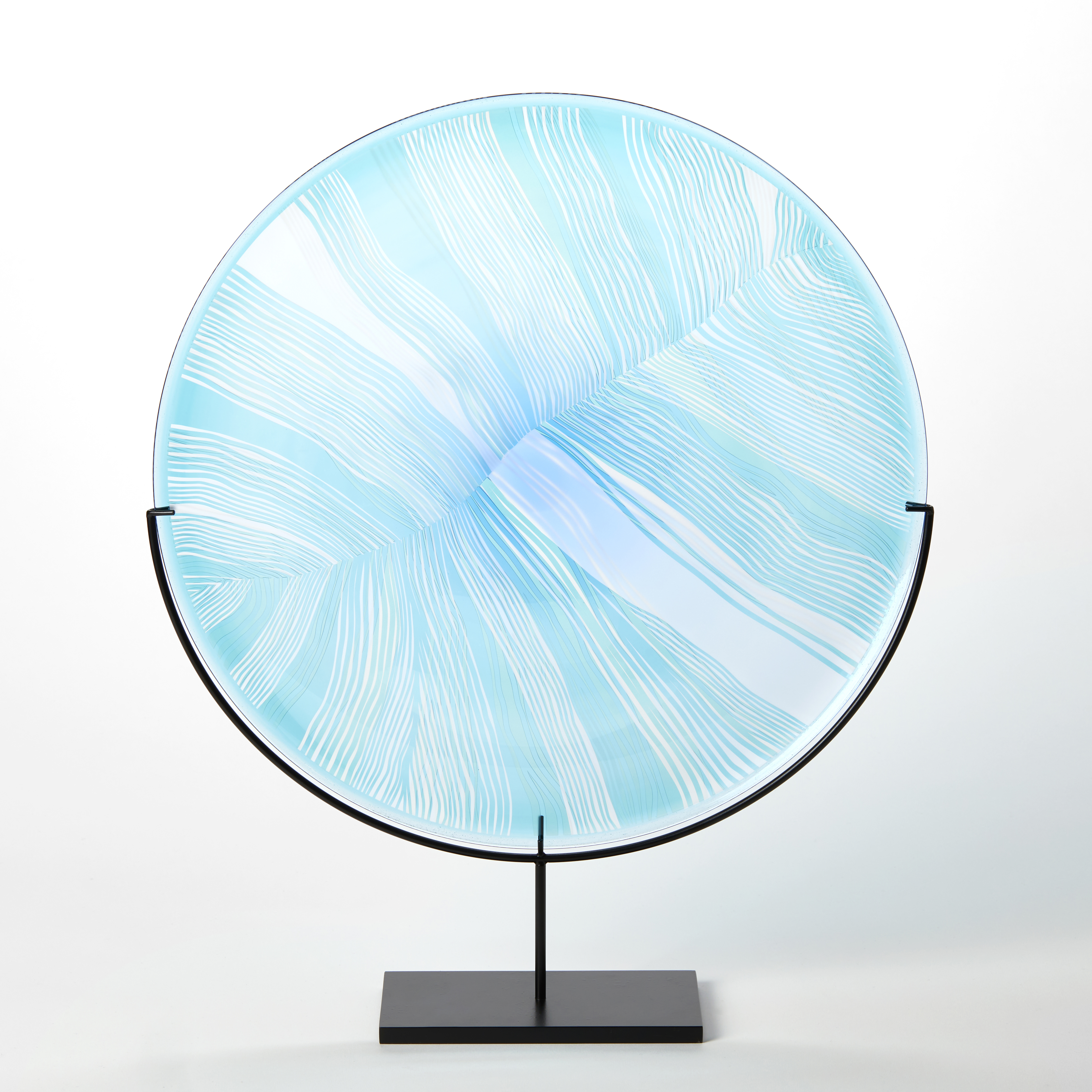 transparent aqua jade and clear glass disc mounted on a black matt stand with cut patterns on the front resembling the magnified details of a birds feather