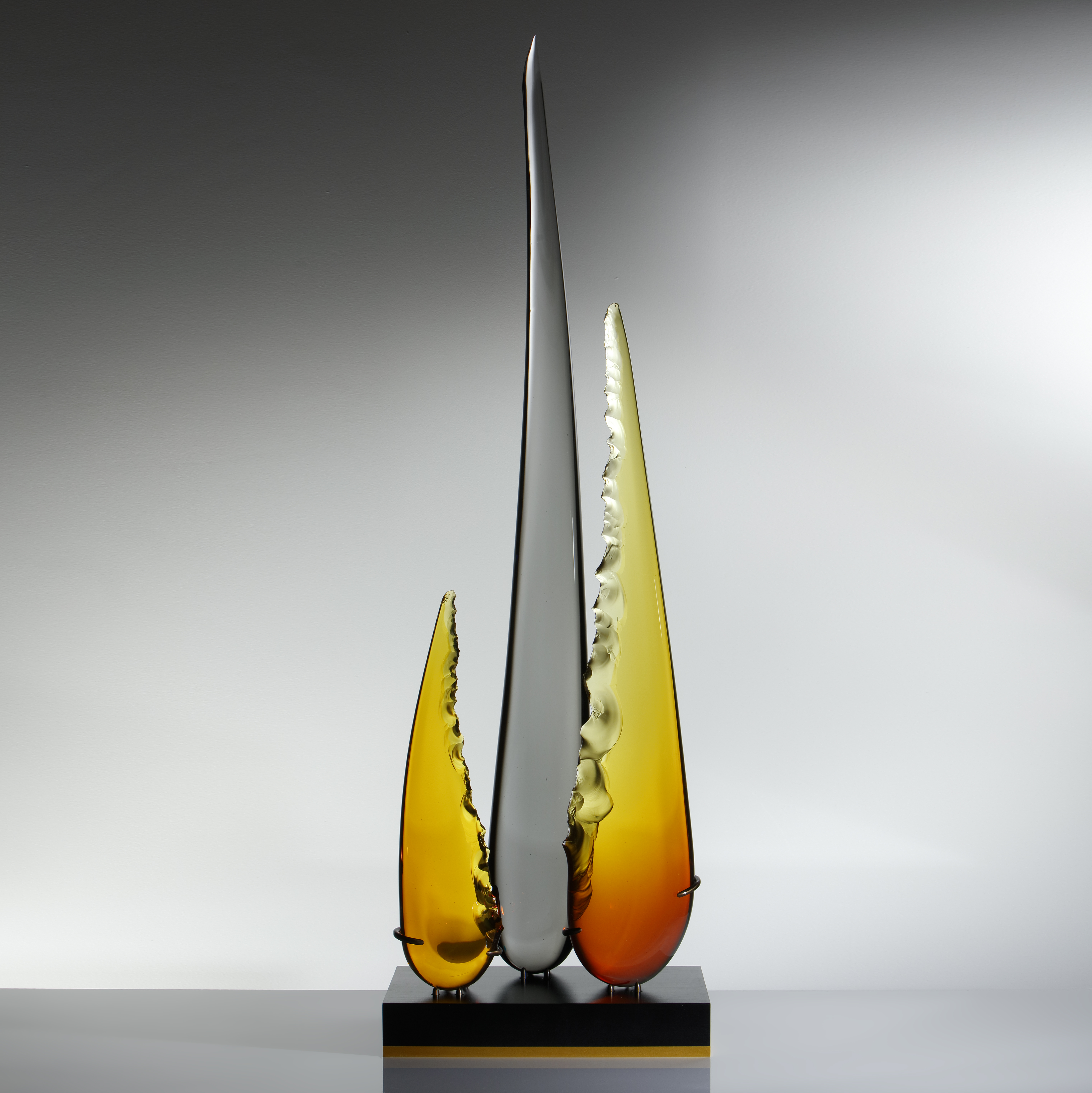 a trio of glass shards one long carbon coloured and centre flanked by two shorter amber and yellow shards resting on black metal base
