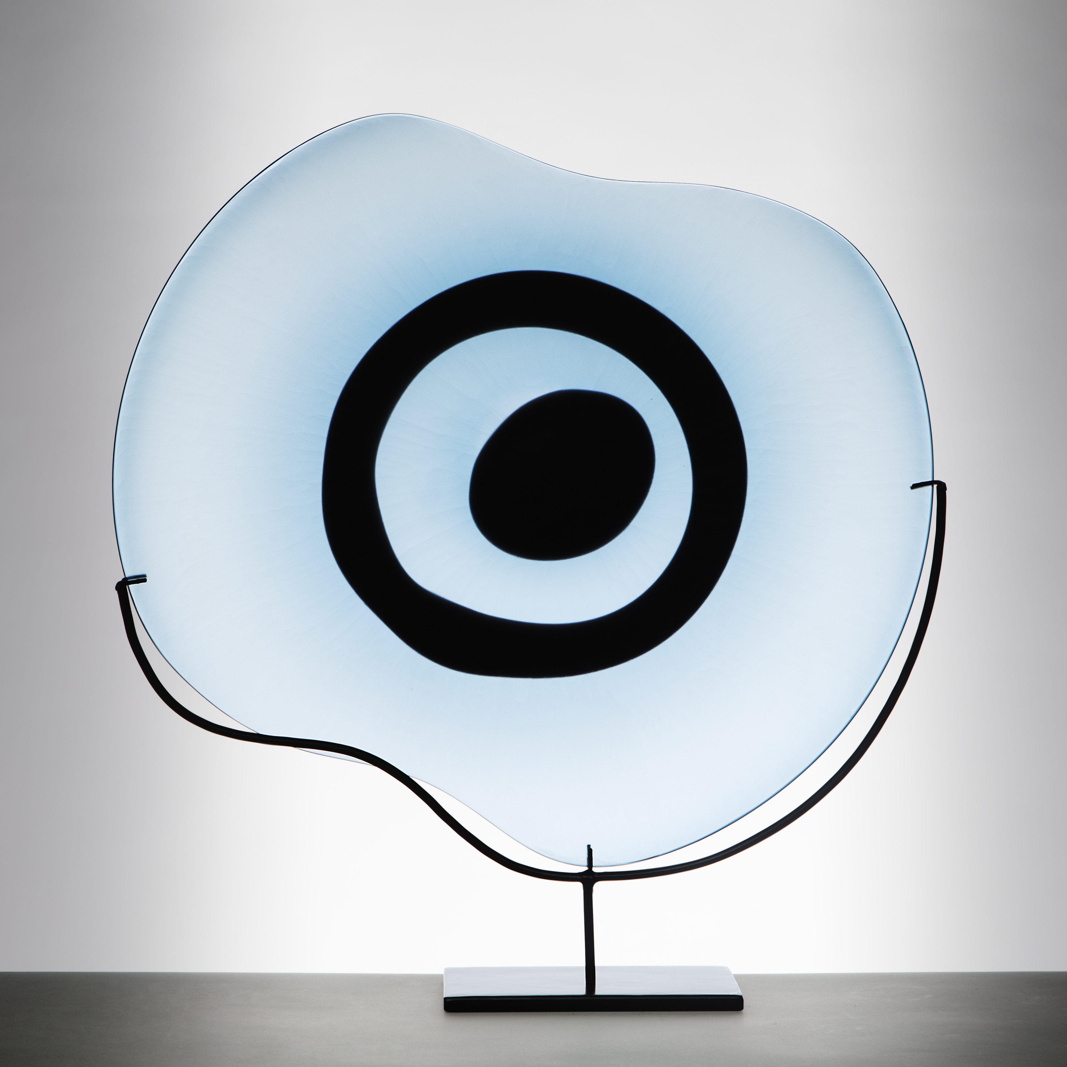 sculpted glass plate in light blue with black eye shape on metal stand