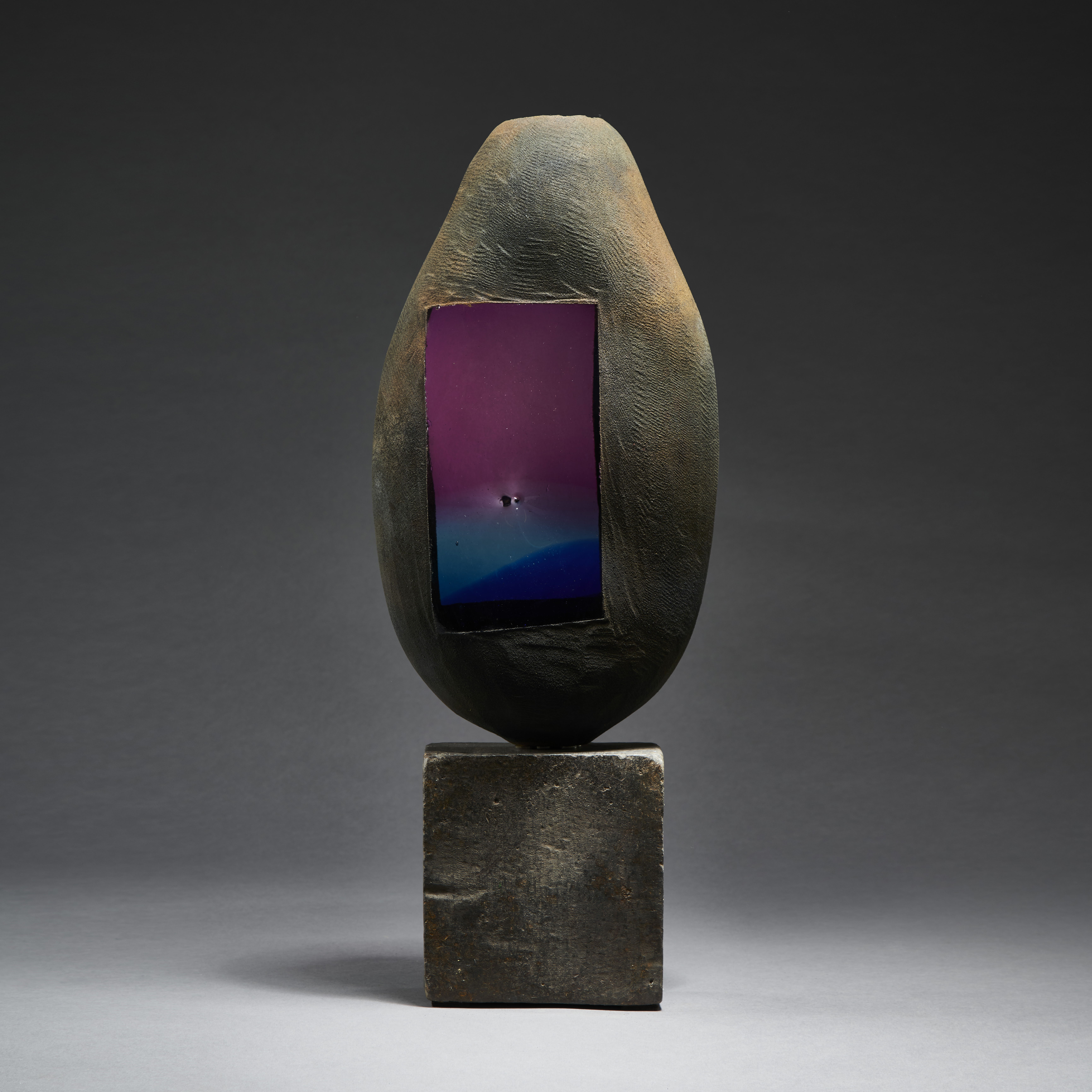 grey and brown rustic battered looking ovoid handmade glass sculpture with faded blue to purple glass window on a beaten metal cube base