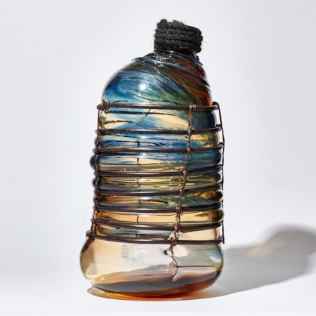 glass sculpture in the shape of an old bottle in blue clear and amber glass in a  copper cage with rope stopper