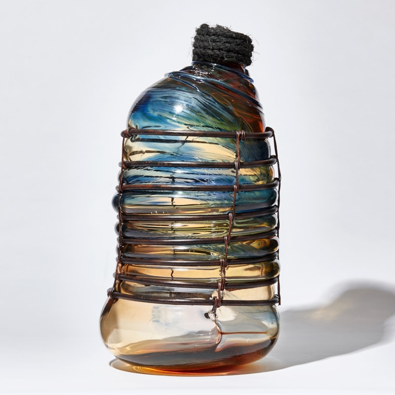 glass sculpture in the shape of an old bottle in blue clear and amber glass in a  copper cage with rope stopper
