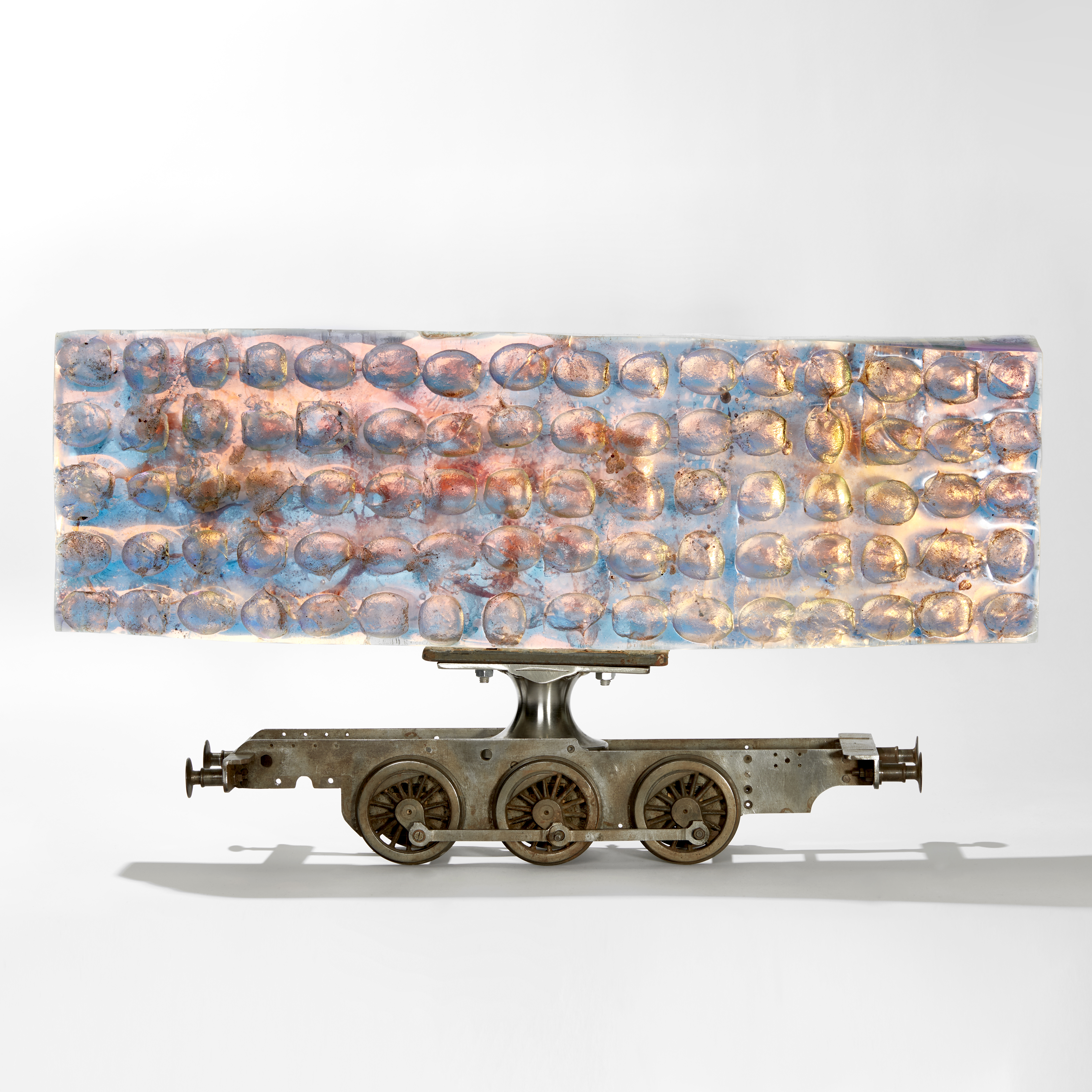 ethereal aqua and jade coloured large rectangular mass of glass with ovoid cutout sections perched on top of a metal scale model train chassis 