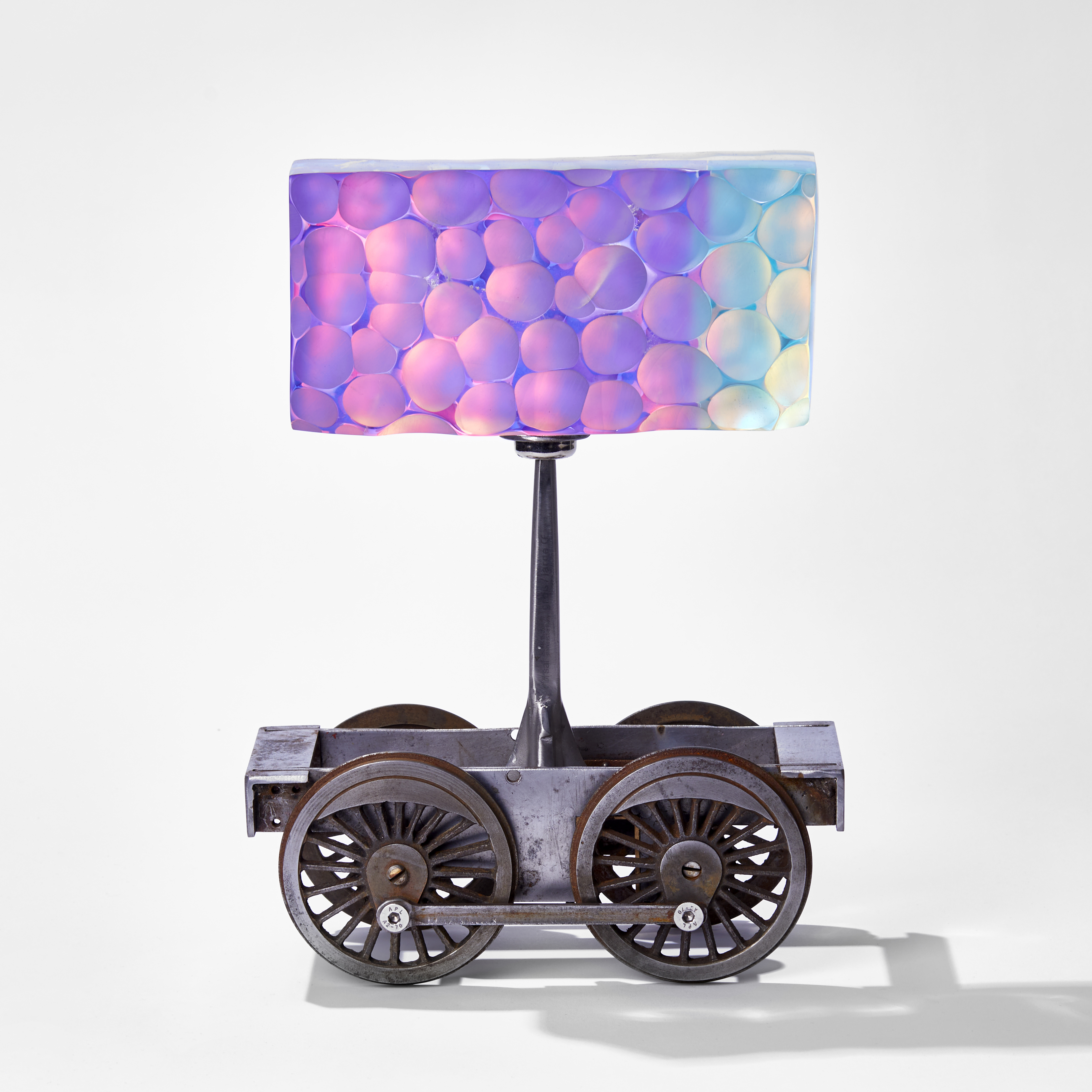 aged metal train chassis with long metal pin above holding up a rectangular dimpled mass of technicoloured glass