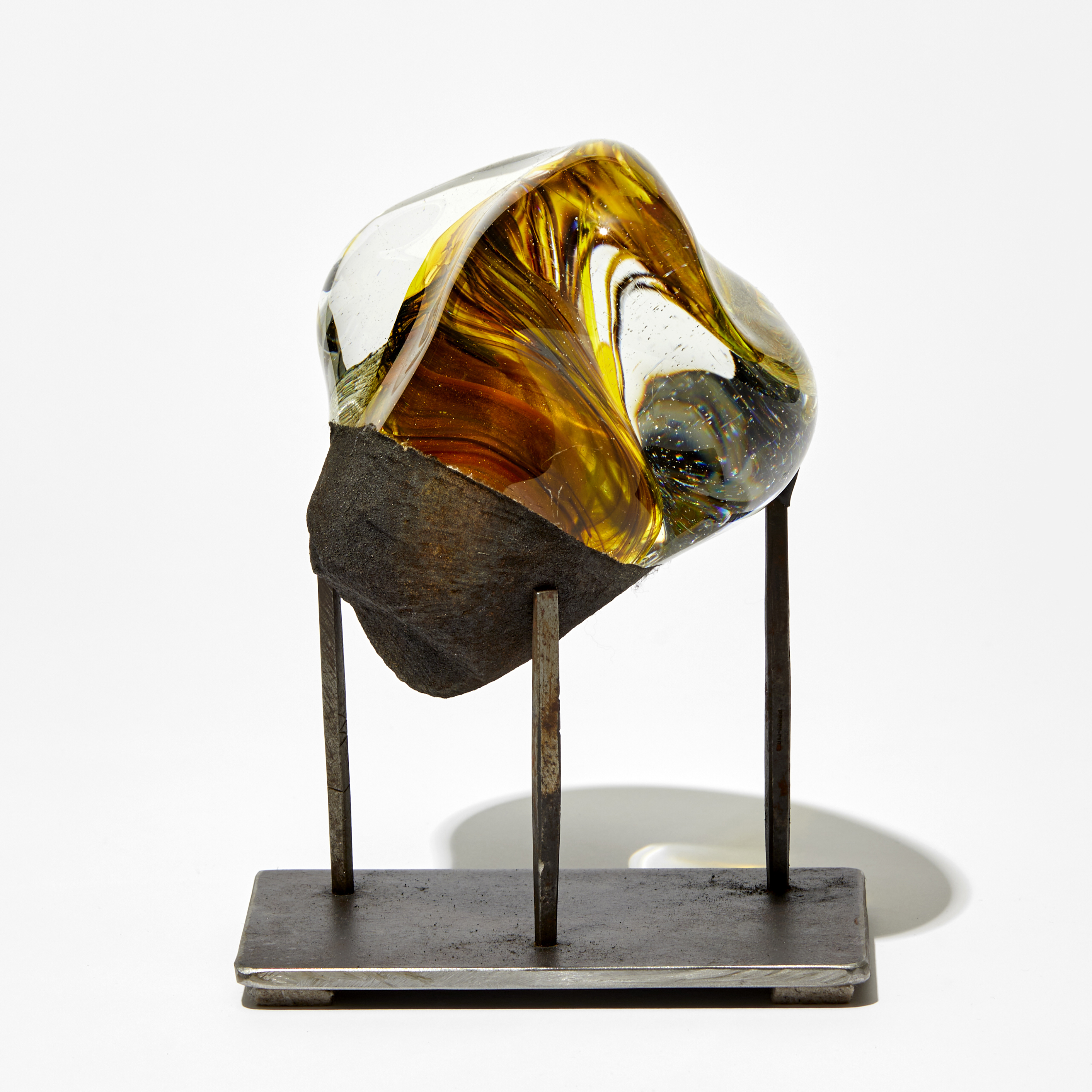 amorphic mass of glass with inner swirling detail in red and yellow perched on a three pronged steel base