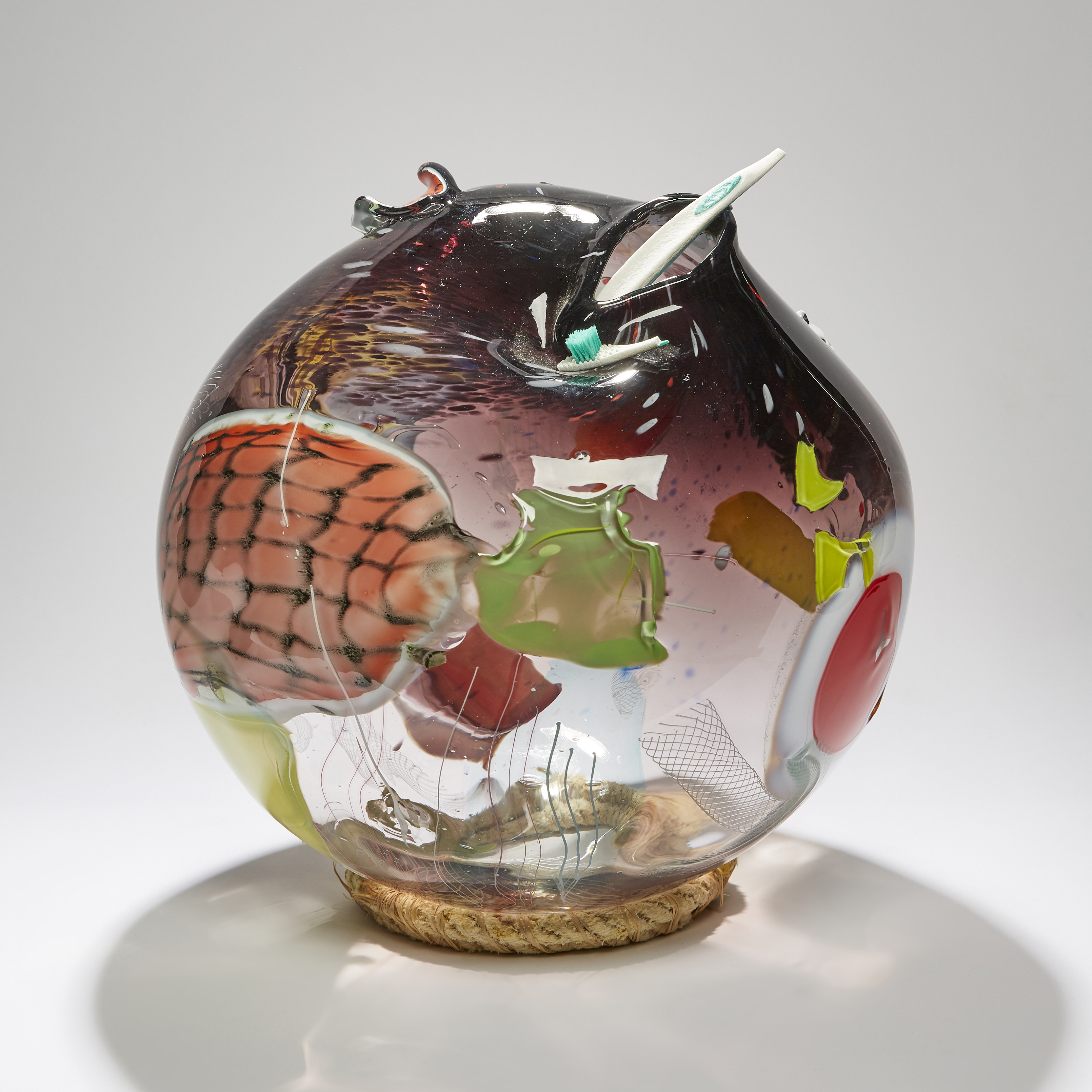rounded vessel in clear and aubergine with latticed patches in yellow coral orange red and white hand made from glass with an ocean plastic toothbrush perched on top