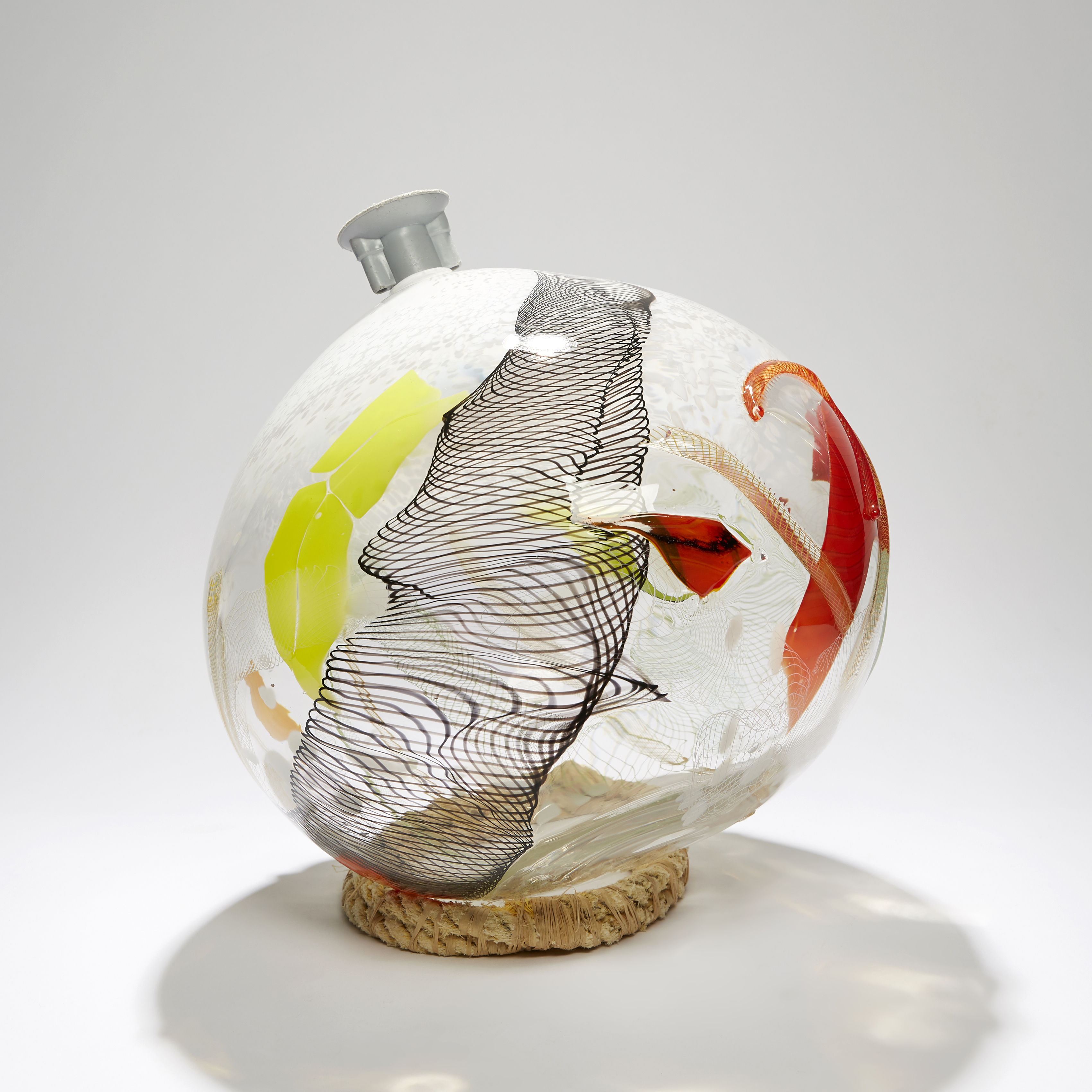 round bottle sculpture in clear with latticed black detail and patches of red yellow and white hand made from glass with ocean plastic fitting at the top