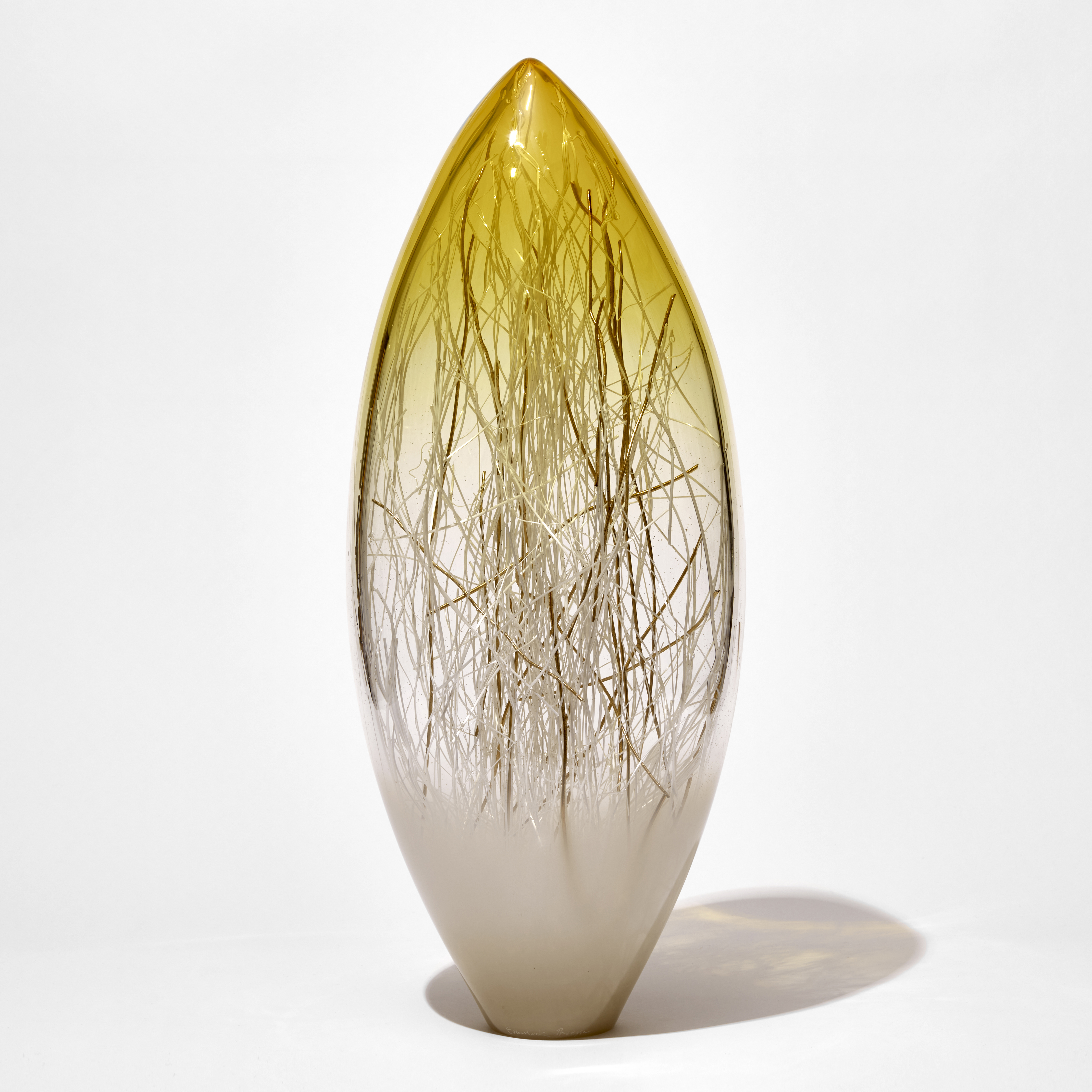 oval pointed sculpture with white base yellow top and clear middle containing thin strands in white and gold hand made from glass